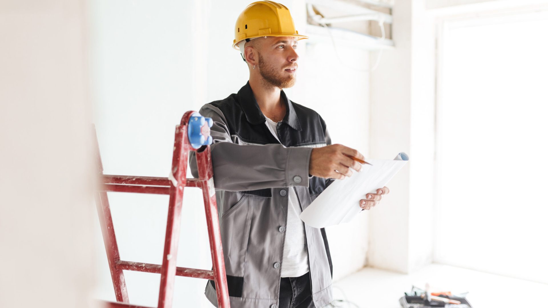 Young engineer in work clothes and yellow hardhat dreamily looking aside holding plan of new apartments in hands with red ladder near in repairing flat